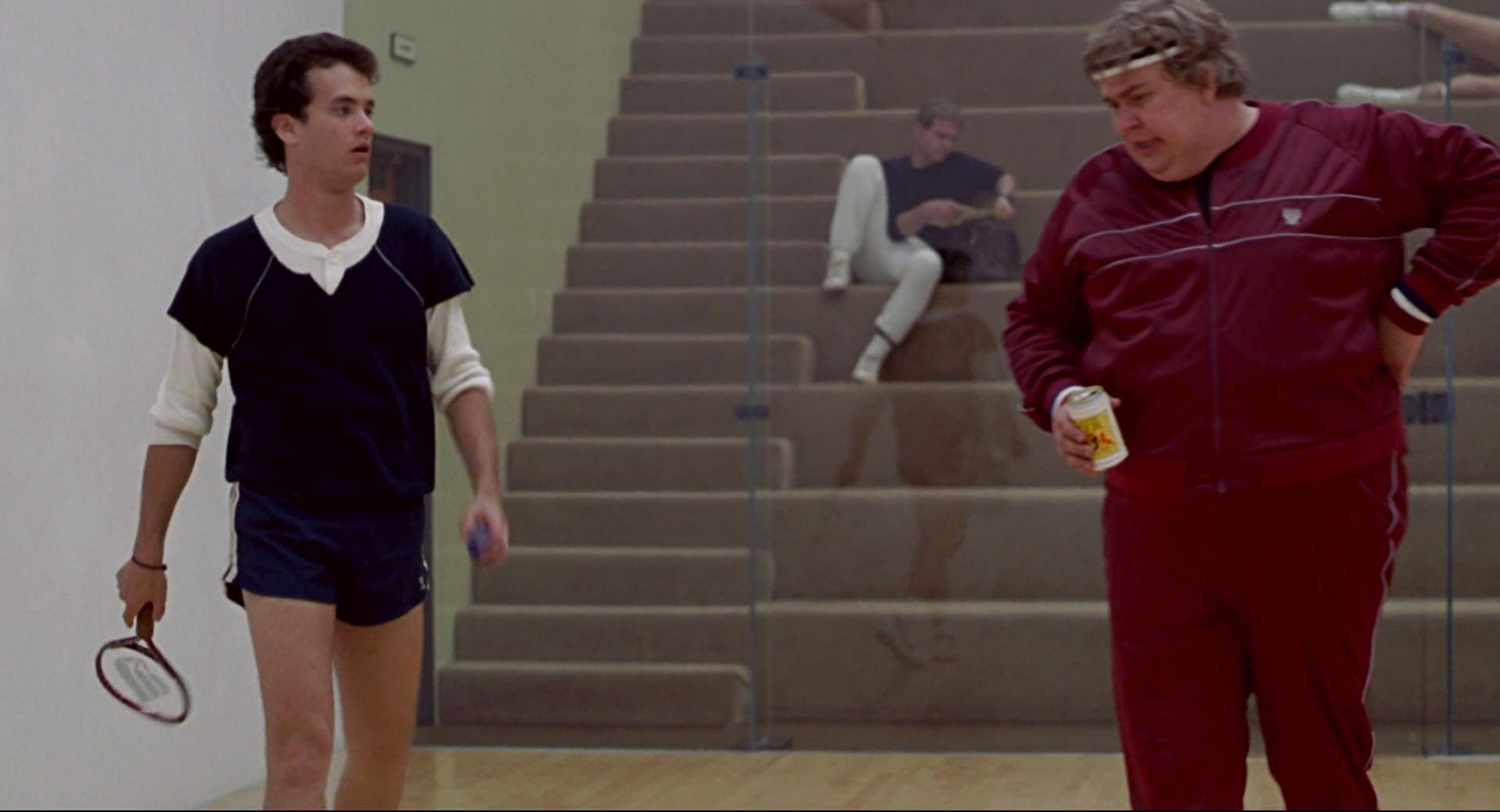 Two men are standing in a gym with one of them holding onto a drink.