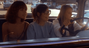 Three women sitting in a car with one holding the steering wheel.